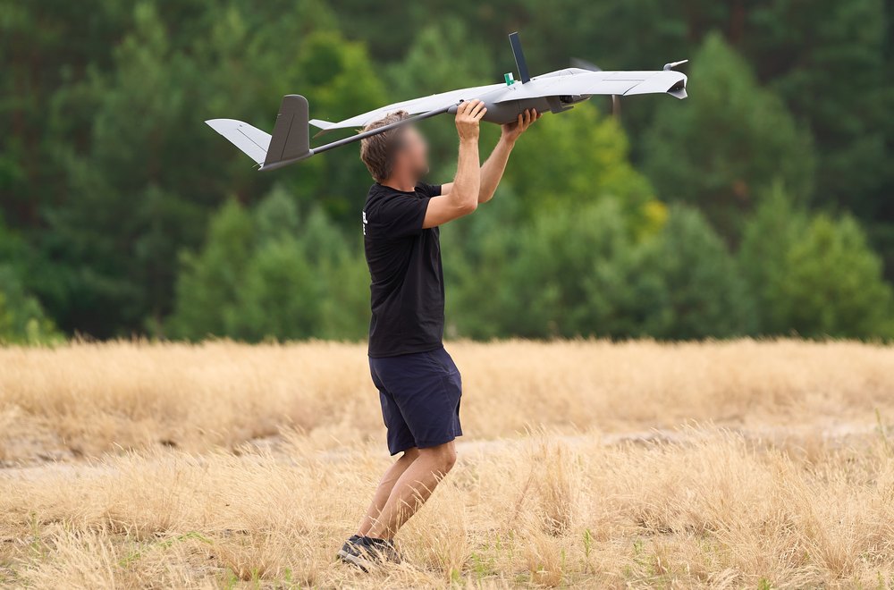 A crew member walking forward across a dry grass field, holding the
                    aircraft above their head at the moment of a hand launch.